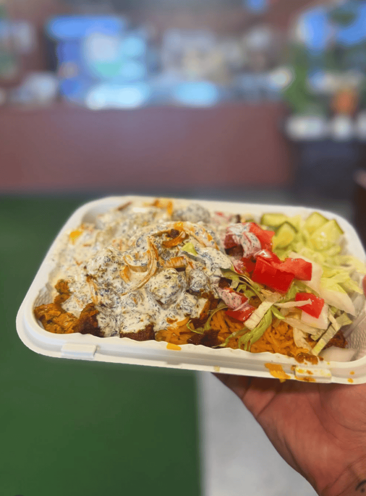 A hand holds a white paper plate of carnival fair food featuring fried chicken, waffle fries, and fresh red peppers at an outdoor event venue.