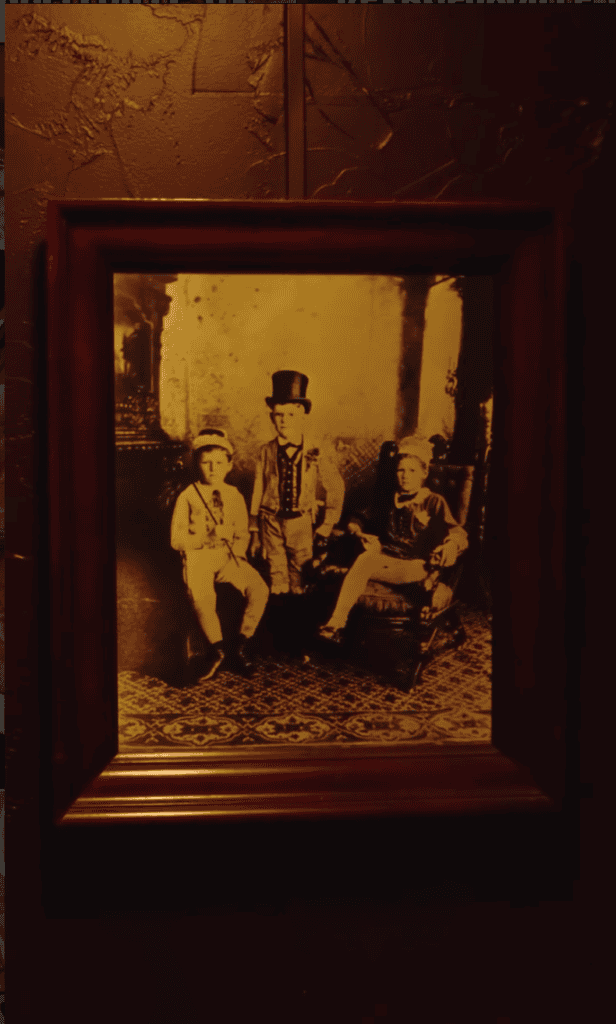 A sepia-toned vintage photograph in a wooden frame hangs on a dark wall, showing three people in period clothing seated indoors.