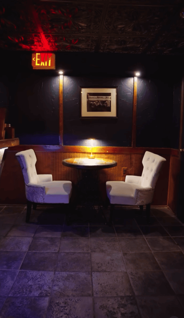 A nighttime outdoor lounge area features two comfortable armchairs arranged around a central fire pit, illuminated by warm amber lighting and framed by dark blue evening tones.