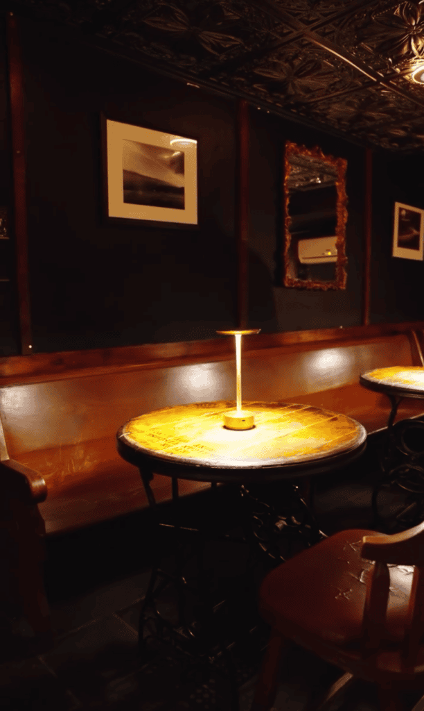 A modern art gallery interior featuring illuminated framed artwork on dark walls, warm accent lighting on a wooden table, and a sophisticated exhibition space.