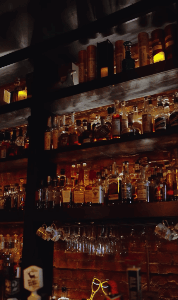 A well-stocked bar shelf displaying numerous bottles of spirits and liquor illuminated by warm golden lighting at night.