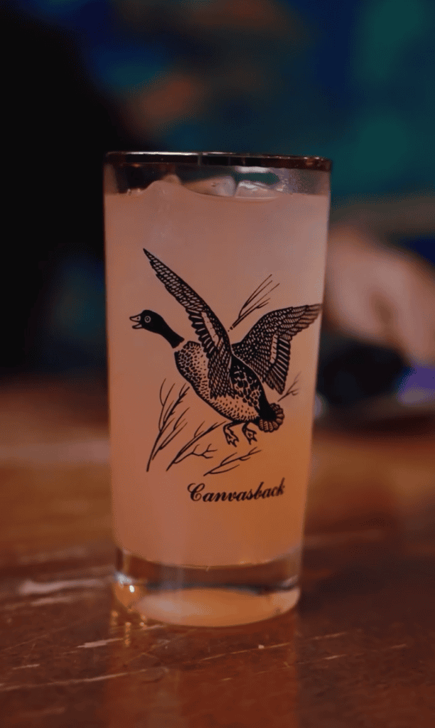 A clear cocktail with a pink hue served in a tall glass, featuring a bird illustration and text reading 'Carmelized' on the glass.