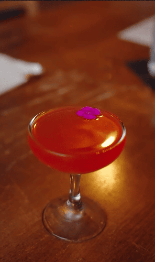 A vibrant red cocktail in a coupe glass with a purple orchid garnish sits on a warm wooden bar surface.