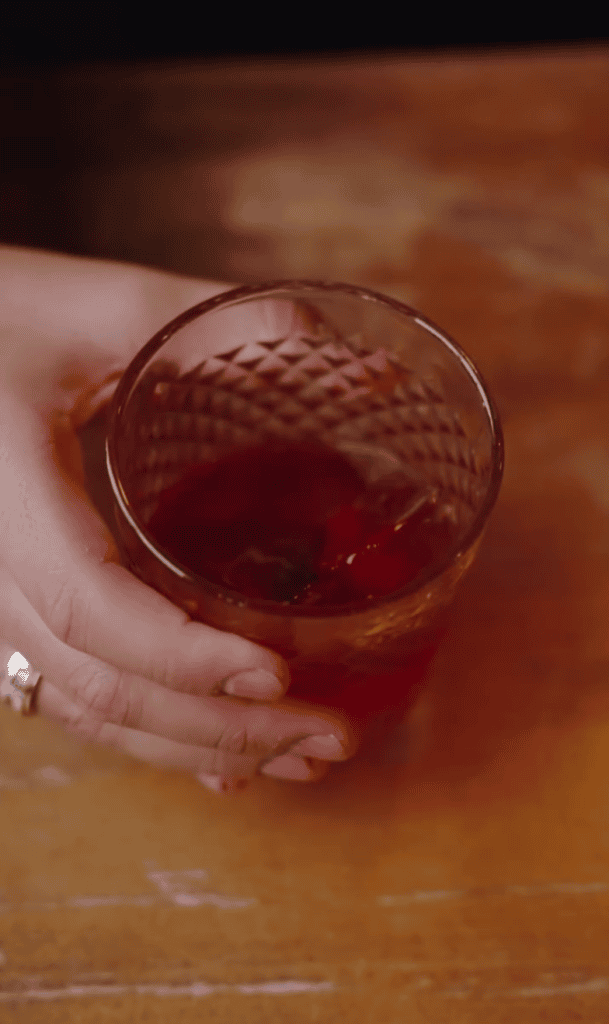 A close-up photograph of a hand holding a glass of amber-colored beverage on a wooden surface.