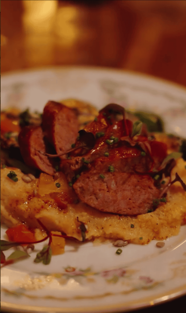 A close-up of a gourmet plated dish featuring sliced sausage and vegetables on creamed polenta served on a white plate.