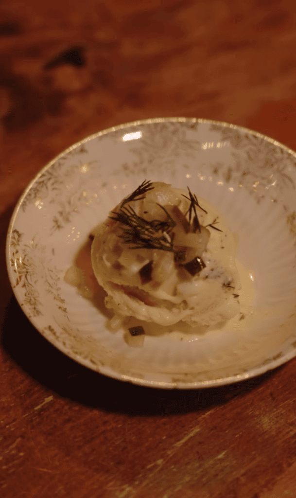 A carefully plated culinary dish with what appears to be a meat or protein component garnished with herbs, served on a ceramic plate against a warm wooden surface.