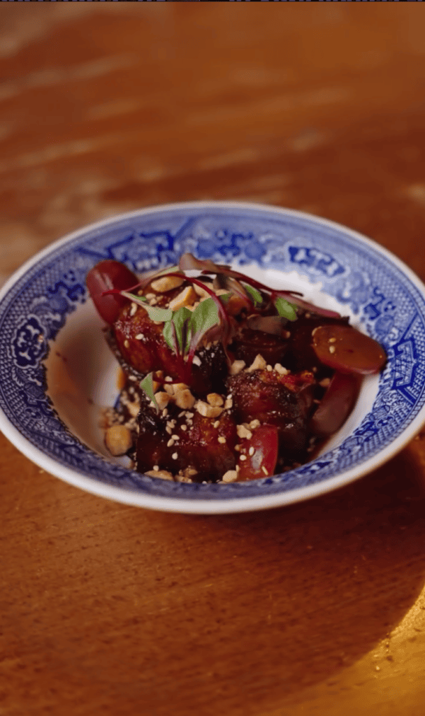 A close-up overhead shot of a braised meat dish garnished with herbs served in a decorative blue and white ceramic bowl on a wooden surface.