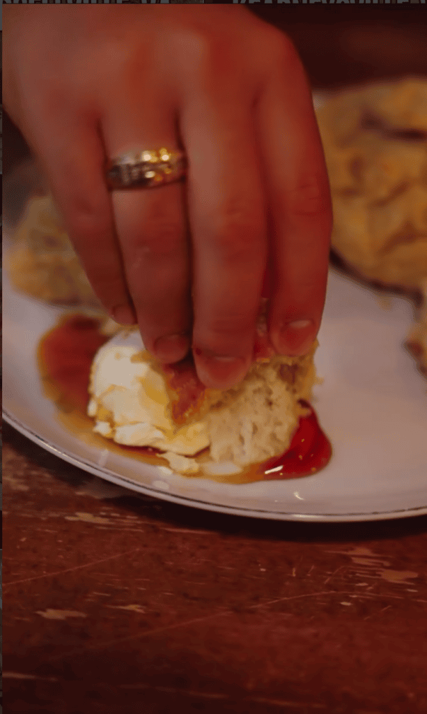 A close-up shot of a hand wearing a gold ring spreading jam on a freshly baked scone on a white plate.