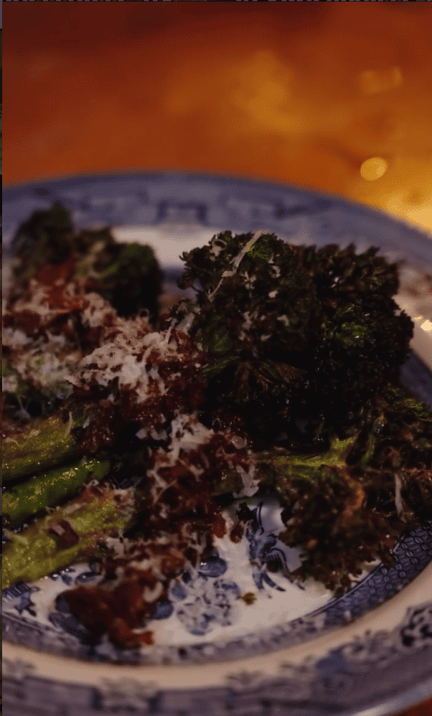 A close-up of an artfully plated salad or vegetable dish with dark greens and garnishes on a white plate, photographed against a dramatic golden-hour sunset sky.