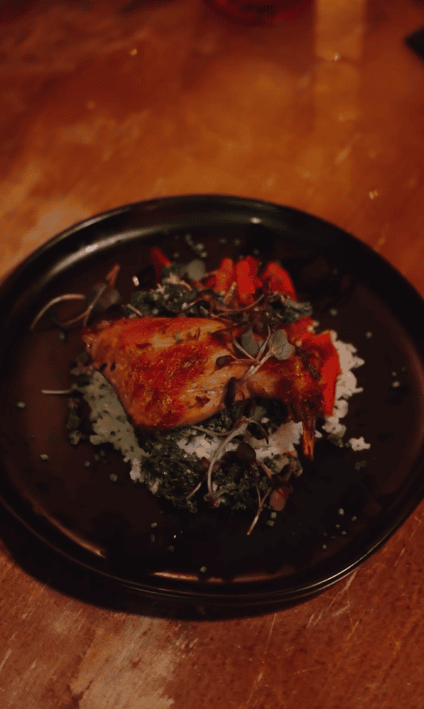 A gourmet plated dish with red/orange protein and herb garnish on a black plate photographed overhead on a wooden surface.