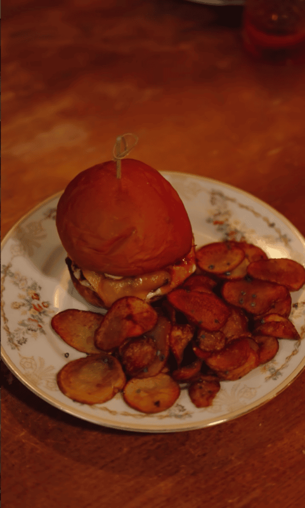 A glossy red candy apple sits on a white plate next to a portion of fried potato slices on a warm-toned surface.