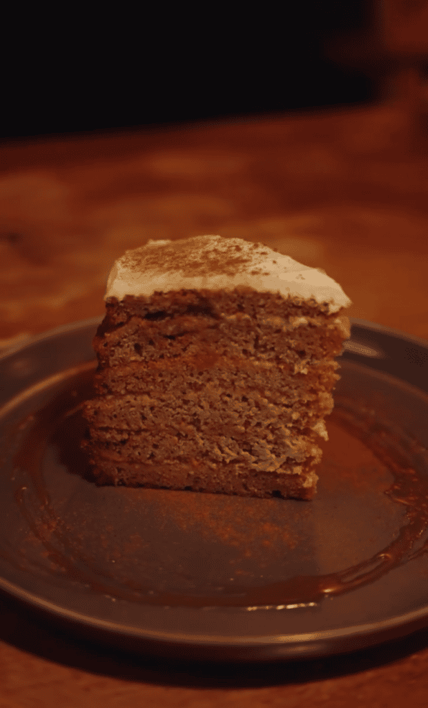 A slice of chocolate cake with frosting sits on a dark plate in warm indoor lighting.