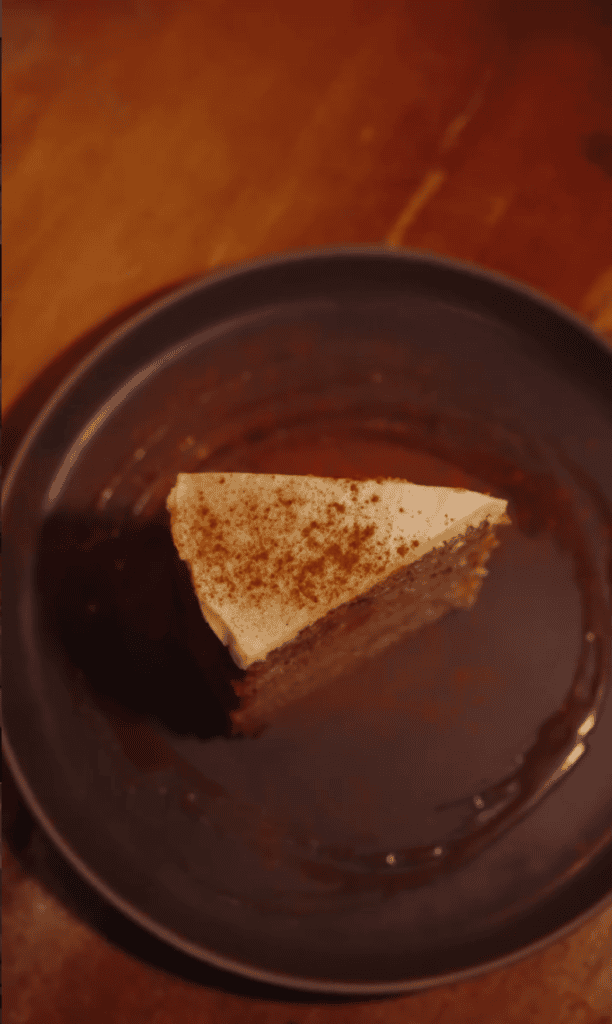A triangular slice of cake with cinnamon-sugar topping sits on a dark plate against a warm brown background.