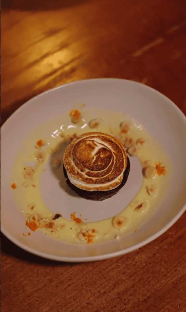 An elegantly plated chocolate dessert with caramel sauce drizzle sits on a white plate against a warm amber-toned surface.