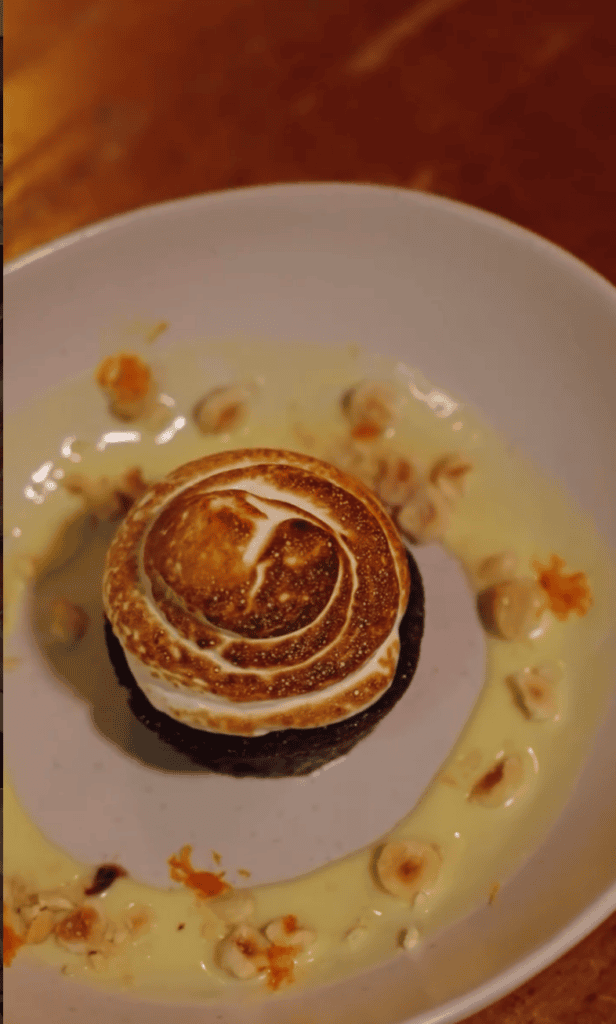 A plated dessert featuring a spiral-shaped pastry with caramel sauce and garnish on a white plate against a warm brown background.