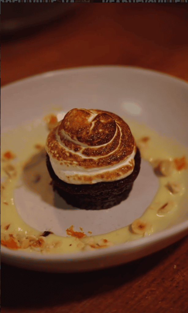 An elegantly plated dessert featuring a chocolate cake base topped with caramel-colored frosting swirl, served on a white plate with sauce drizzle against a wooden surface.