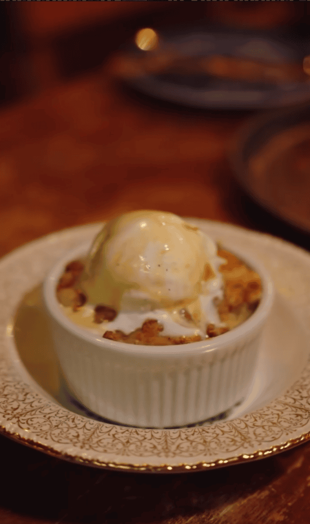 A plated individual fruit dessert in a white ramekin topped with whipped cream and caramelized fruit garnish, presented on a textured placemat.