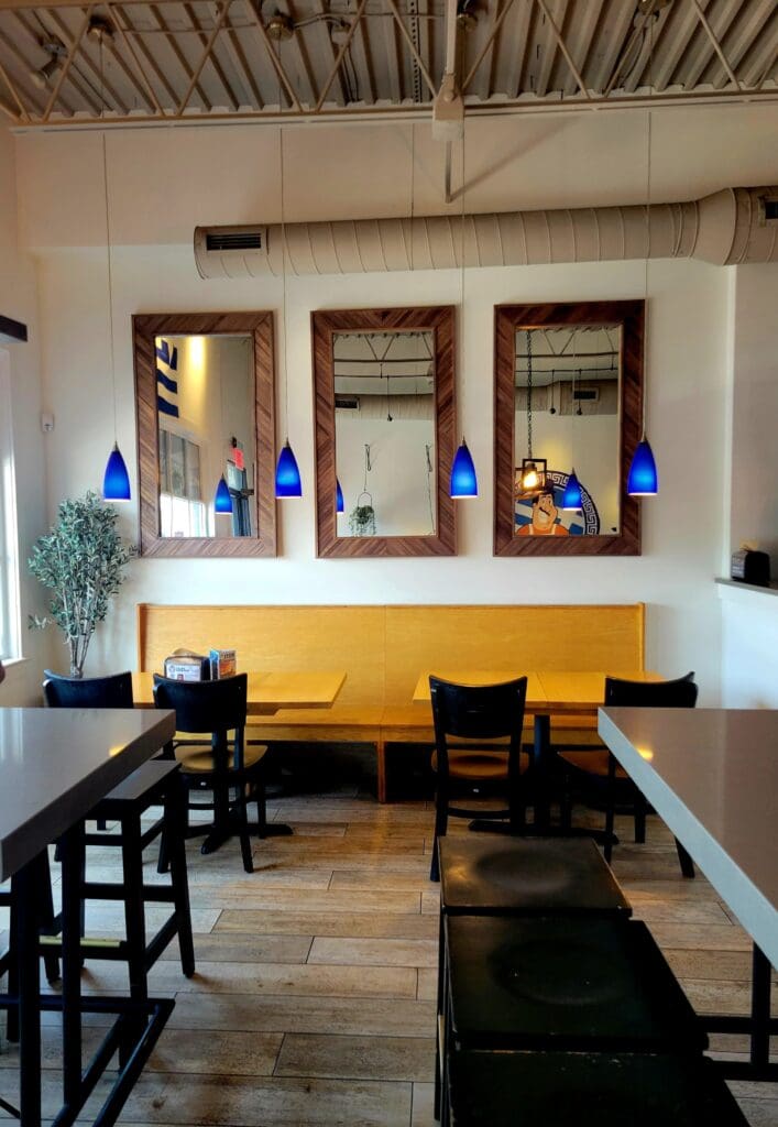 A modern restaurant interior featuring a bright yellow banquette seating area with black chairs, blue pendant lights, and framed artwork on the walls.