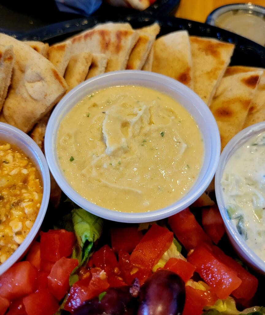 An overhead view of Mediterranean appetizers featuring bowls of hummus and dips surrounded by pita bread, fresh red peppers, and other vegetables on a wooden platter.
