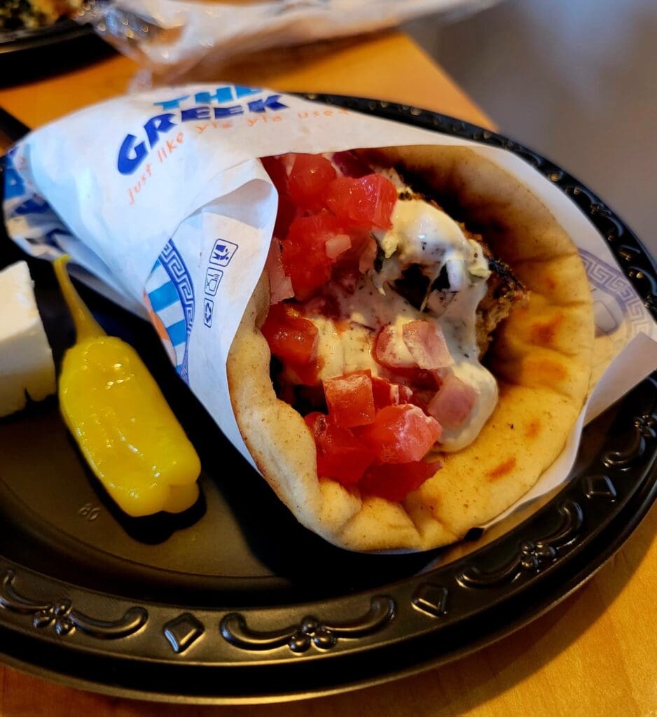 A close-up of a gourmet hot dog topped with red peppers and other toppings, served on a decorative black plate with a pickle and wrapped in paper.