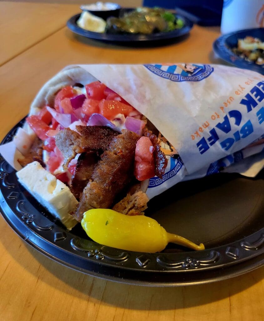 A close-up of a freshly grilled meat sandwich topped with diced tomatoes and onions, wrapped in paper on a decorative blue and white plate with a yellow pepper.