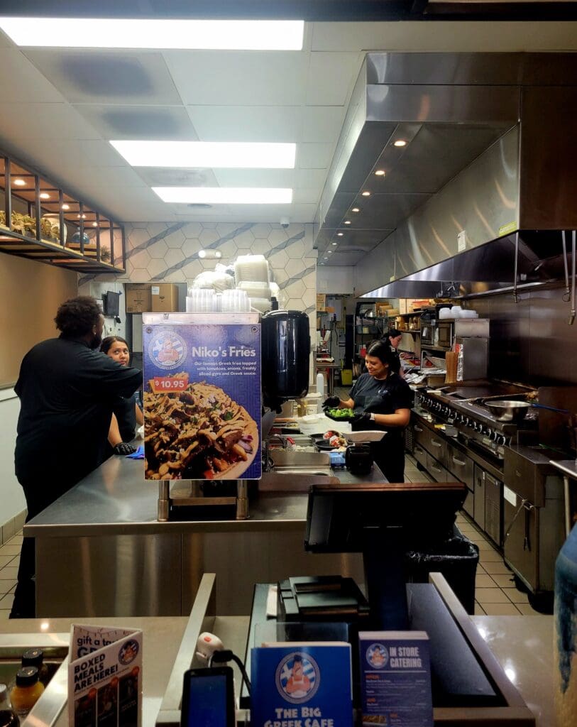 A busy commercial kitchen with staff working at various stations, featuring stainless steel equipment and a food display visible in the foreground.