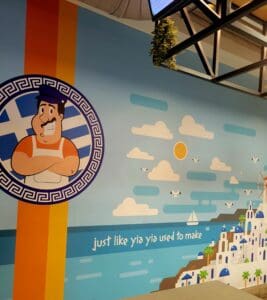 A themed indoor venue features a large Hercules-inspired mural with Mediterranean architectural elements, clouds, and motivational text against a blue sky background.