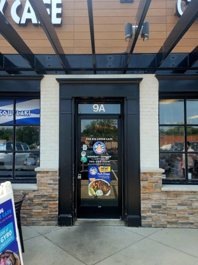 A commercial storefront with a black-framed glass entrance door and brick facade under a blue beam overhang in daylight.
