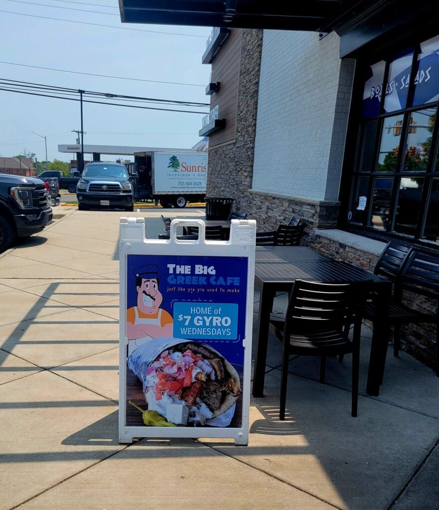 A roadside gyro restaurant storefront with a blue and white promotional sign displaying food imagery, located in a strip mall parking lot during daytime.