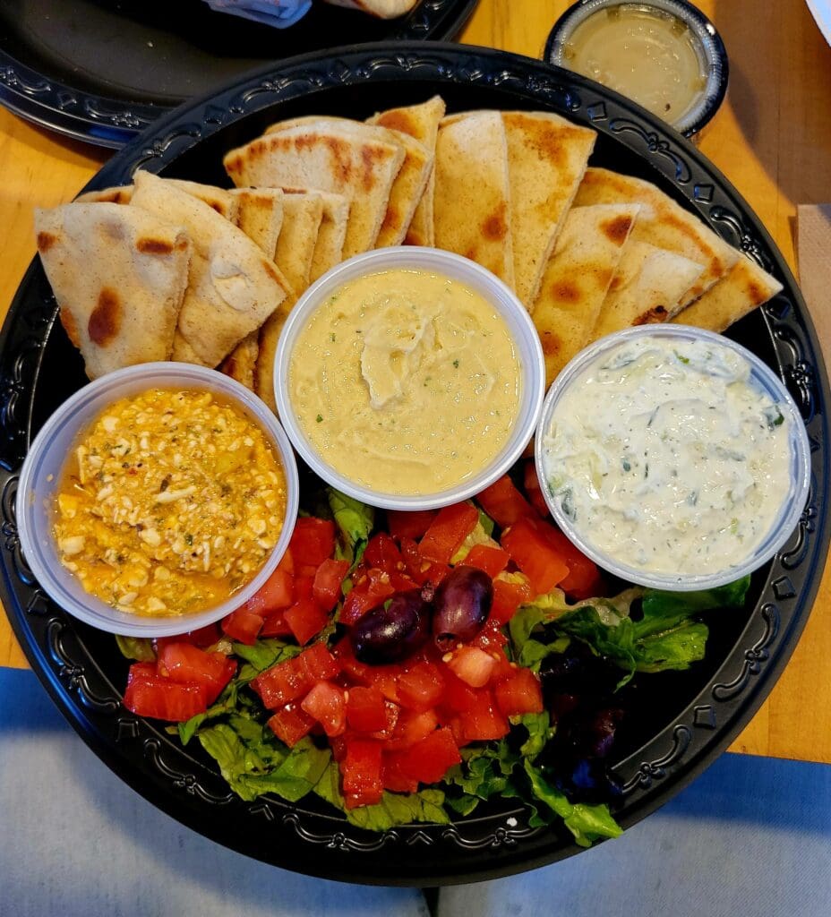 An overhead view of a black serving platter filled with various dips, hummus, pita chips, and fresh red peppers arranged for entertaining or dining.