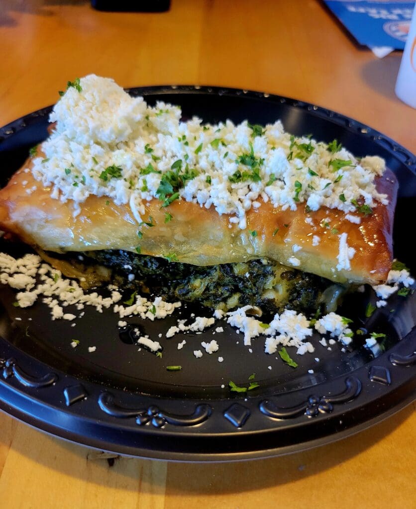 A close-up overhead view of a plated enchilada topped with melted white cheese and green garnish on a black plate in an indoor dining setting.