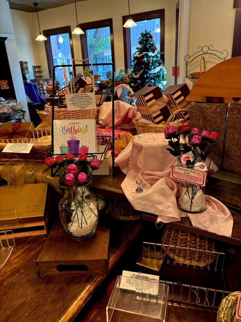 An elegantly decorated interior venue displays multiple floral arrangements, a decorated Christmas tree, and holiday decorations arranged on wooden furniture with windows visible in the background.