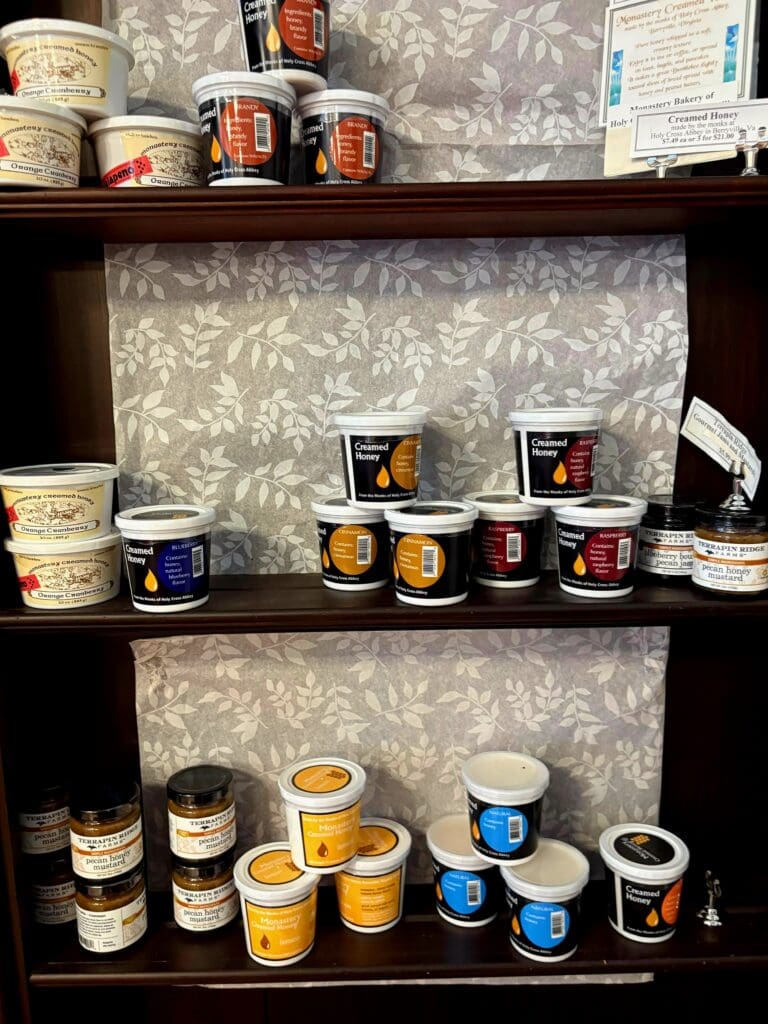 A well-organized pantry shelf displaying various canned goods, jars of preserves, and stored food items arranged on three wooden shelves against a patterned wallpaper backdrop.