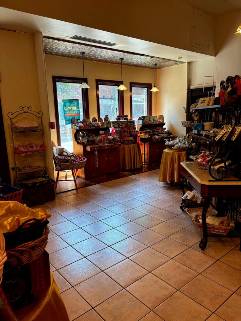 A warm-toned retail or workshop interior displaying merchandise on wooden displays and clothing racks along a tiled floor.
