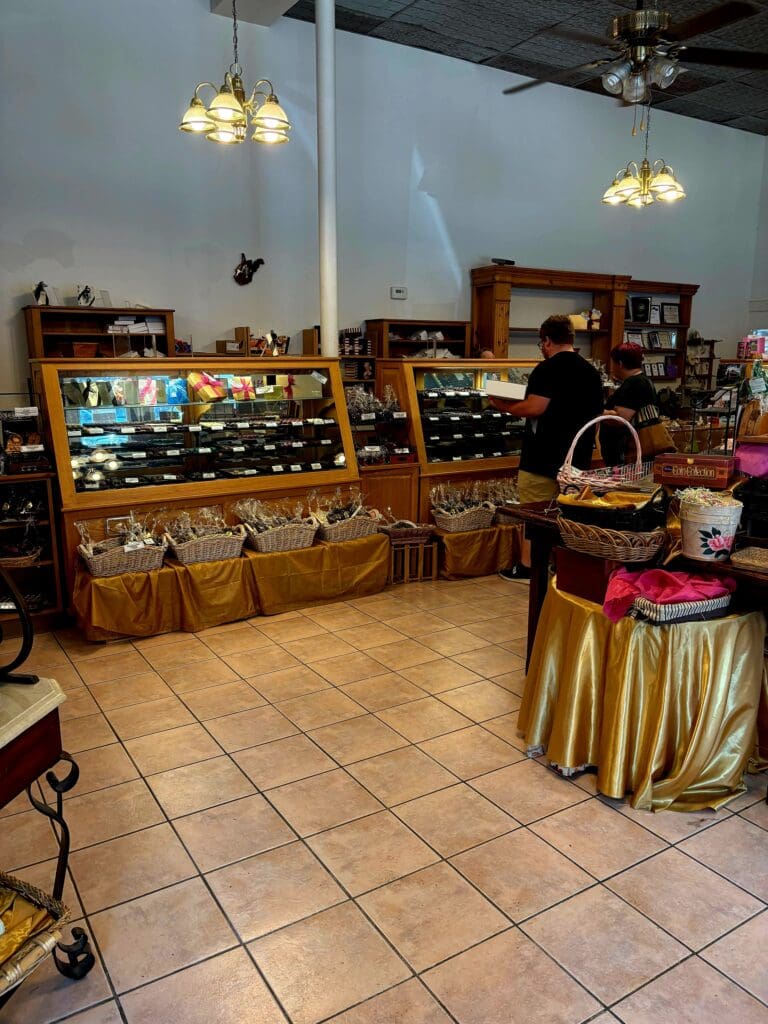 An indoor vendor booth at a marketplace or fair displays merchandise in glass cases and under gold-draped tables with hanging lights overhead.