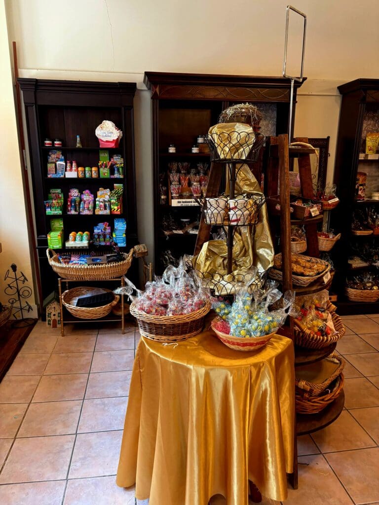 A religious gift shop or shrine displays elaborate floral arrangements on a gold-draped table in front of a vending machine and shelving with religious items.