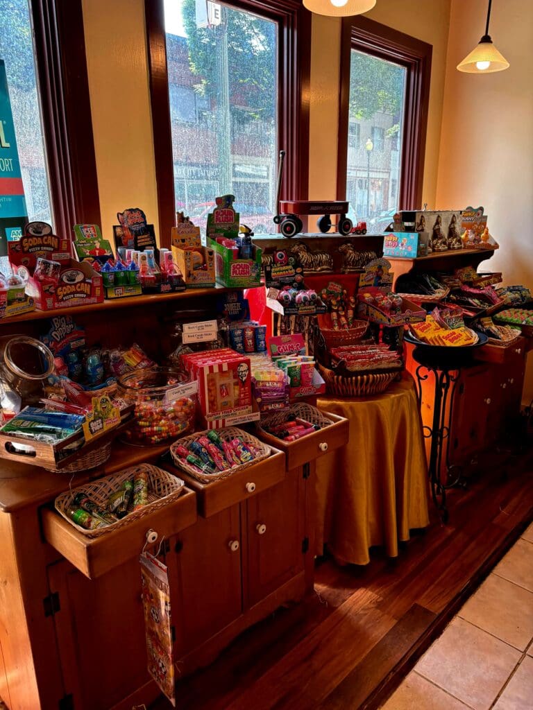 An indoor holiday craft fair or market display featuring multiple tables draped in red fabric showcasing handmade goods and decorative items near large windows.