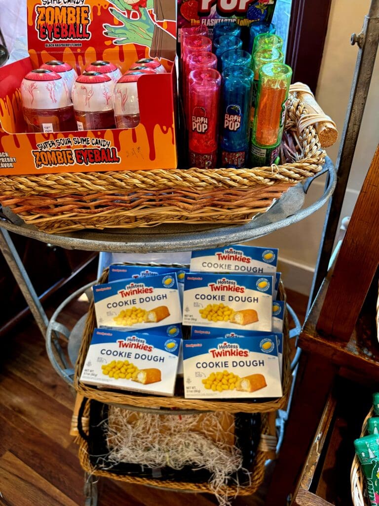 A tiered display cart showcasing various packaged food and beverage products with a wicker basket on top, arranged in an indoor retail or vendor setting.