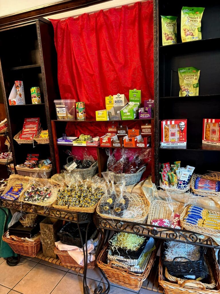 A carnival or fair food vendor display with shelving units stocked with colorful snacks, popcorn, candy, and beverages behind a prominent red curtain backdrop.
