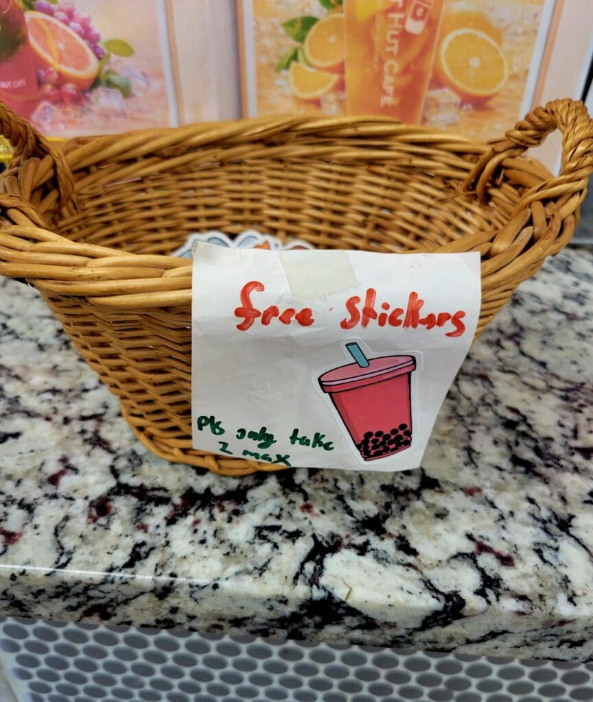 A handmade sticker package labeled 'Soo Stickers' with a cute drink illustration sits in a wicker basket on a granite countertop.