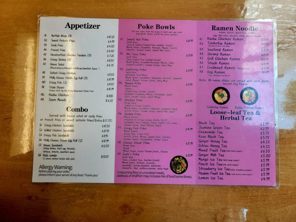A restaurant menu with three tri-fold pages displaying appetizers, poke bowls, ramen noodles, and desserts with prices on a wooden table surface.