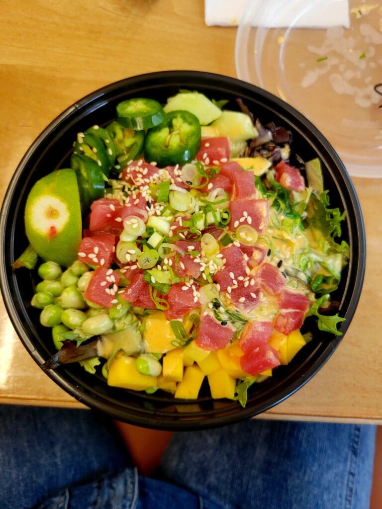 An overhead view of a fresh vegetable salad in a black bowl with green peppers, tomatoes, corn, and leafy greens on a wooden surface.