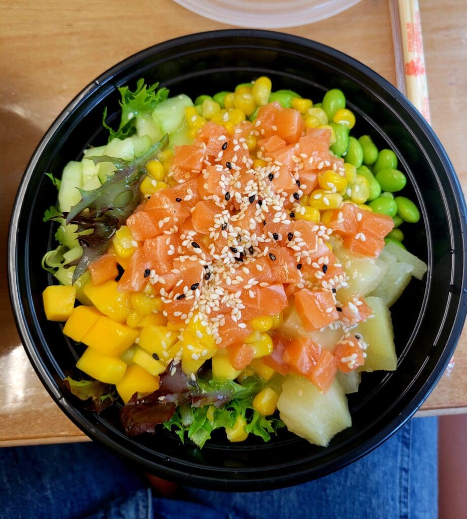 An overhead view of a colorful poke bowl with salmon, corn, cucumber, and other vegetables topped with sesame seeds in a black container.