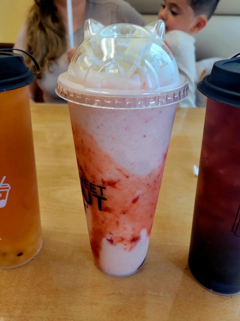 A close-up of a bubble tea drink with pink-tinted liquid and black boba pearls in a clear plastic cup with dome lid, displayed on a counter with other beverages in the background.