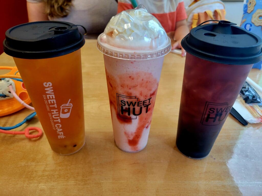 Three colorful boba tea drinks in plastic cups arranged on a wooden table, featuring orange, pink, and dark purple beverages with dome lids.