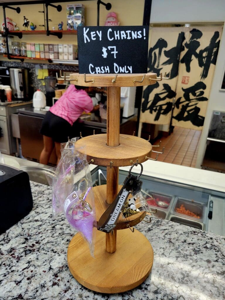 A wooden tiered display stand showcasing key chains and merchandise in a craft workshop or retail setting, with a hand-written chalkboard sign identifying the products.