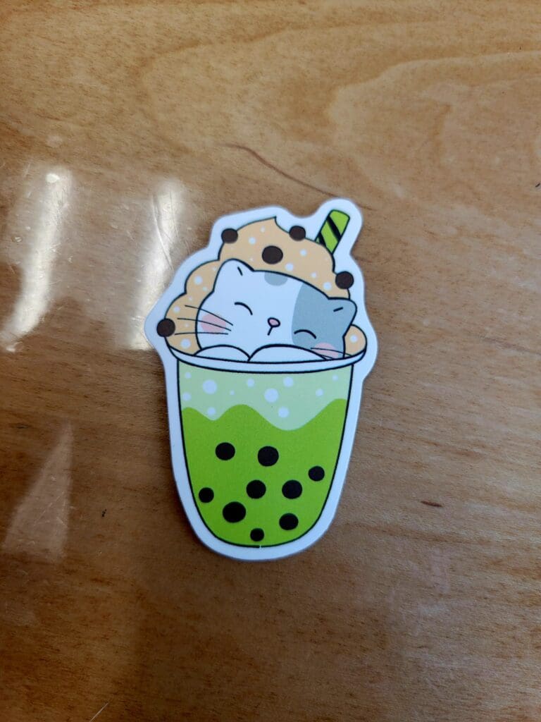 A cute cartoon bubble tea keychain charm with a smiling face and green drink with boba pearls, photographed on a wooden surface.