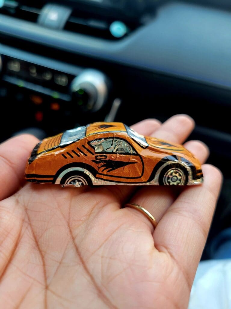 A close-up of a hand holding an orange die-cast toy car inside a vehicle, with the dashboard visible in the blurred background.