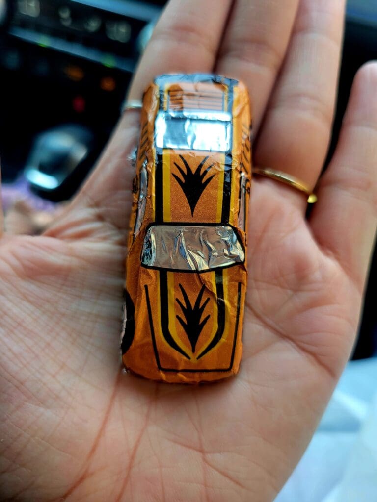 A close-up of a hand holding a vintage orange toy car with black flame details against a car interior background.