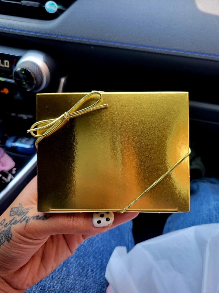 A person holds a small gold gift box with a rope tie inside a vehicle, presenting it as an unwrapped present.